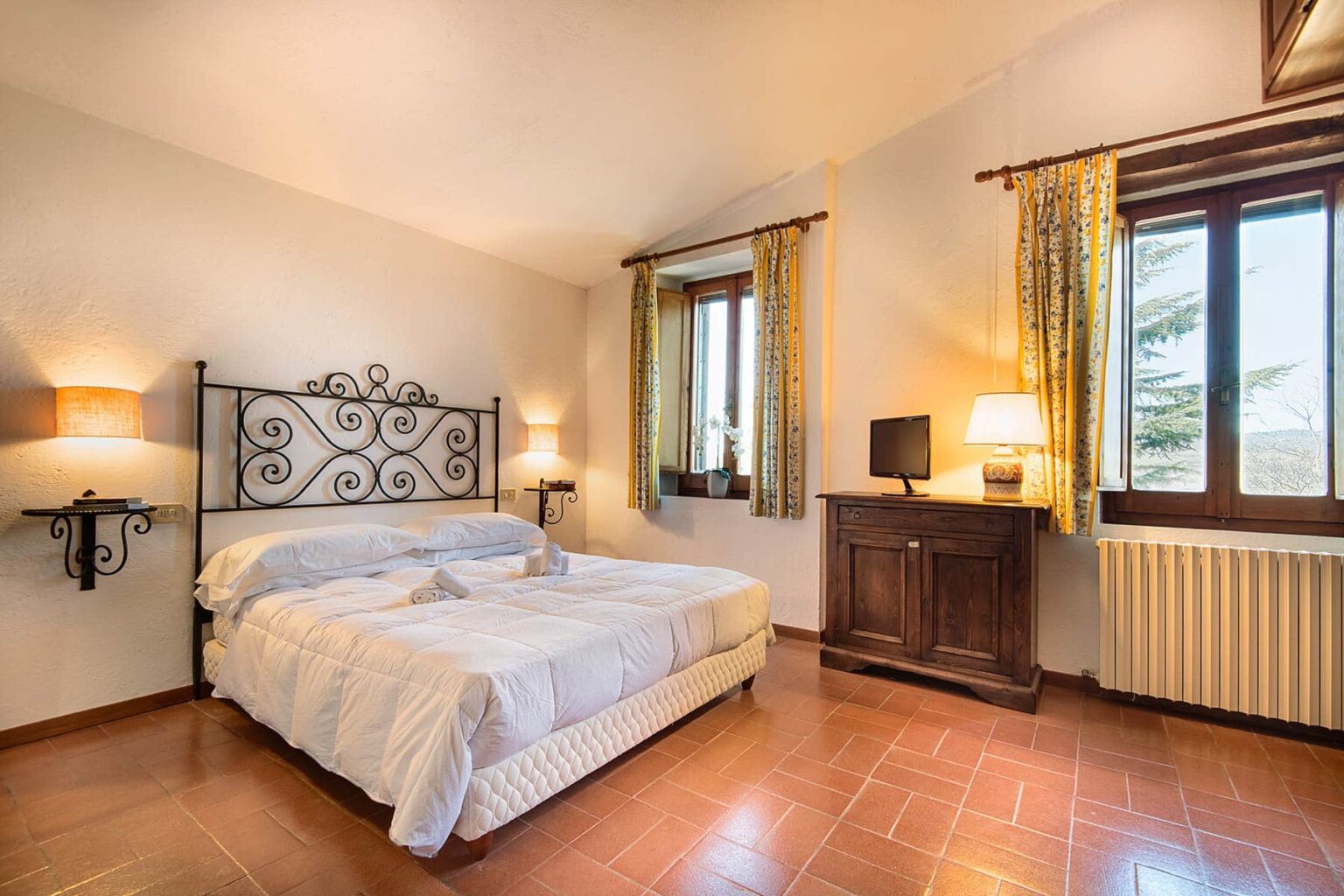 Cozy bedroom featuring a double bed with decorative headboard, wooden furniture, and large windows with yellow curtains, creating a bright and inviting atmosphere. | Only on SopranoVillas