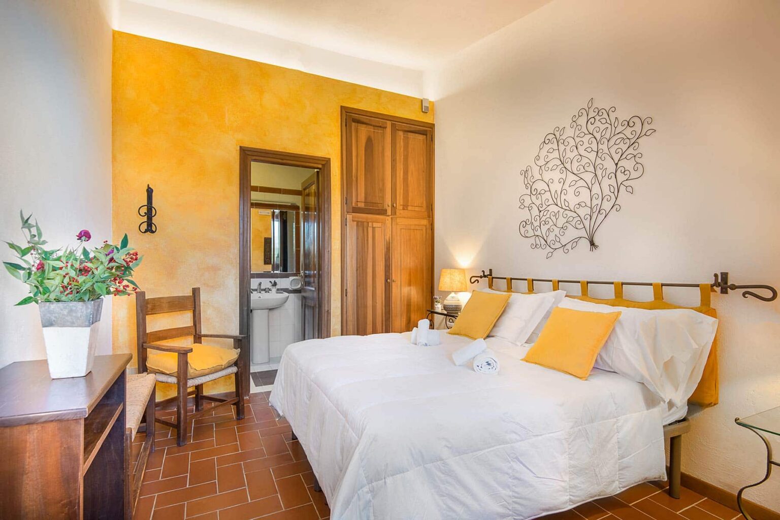 Cozy bedroom with a double bed adorned with yellow pillows, a wooden chair, and a bedside table. The walls are painted in warm yellow tones, and a decorative leaf design adds a touch of elegance. A small bathroom is visible through the open door, enhancing the room's functionality. | Only on SopranoVillas