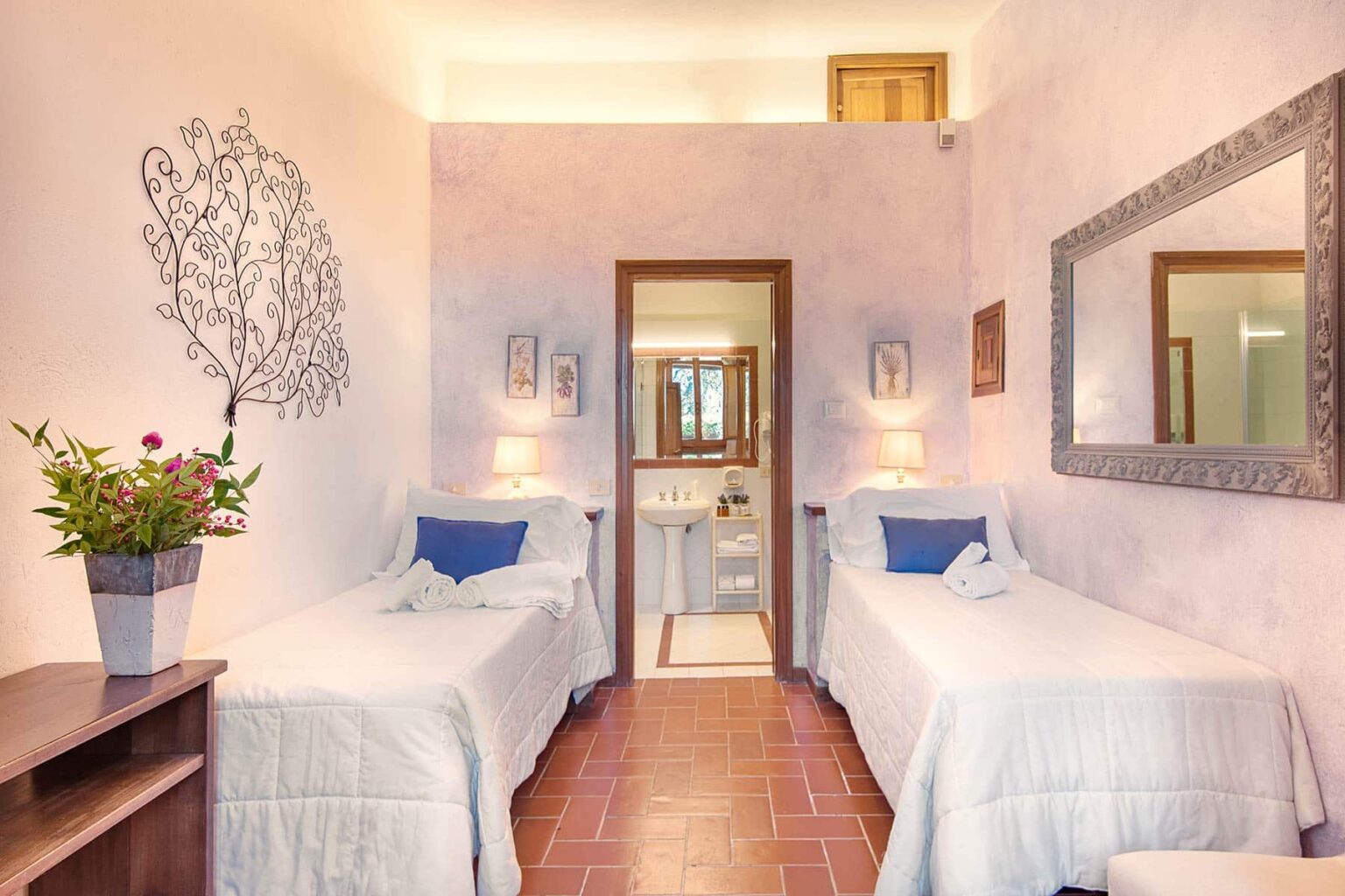 Cozy guest room featuring two twin beds with white bedding, a decorative wall art of a tree, a wooden shelf with a flower pot, and a bathroom entrance in the background. Ideal for travelers seeking a comfortable and inviting stay. | Only on SopranoVillas