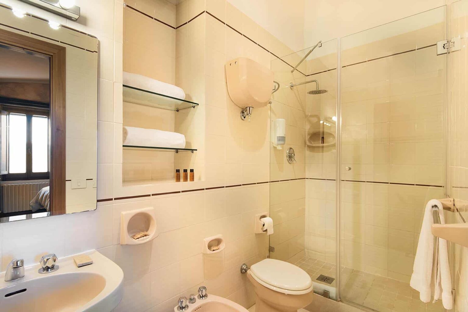 Modern bathroom with glass shower, toilet, sink, and shelves holding towels and toiletries, featuring neutral tones and natural light. | Only on SopranoVillas