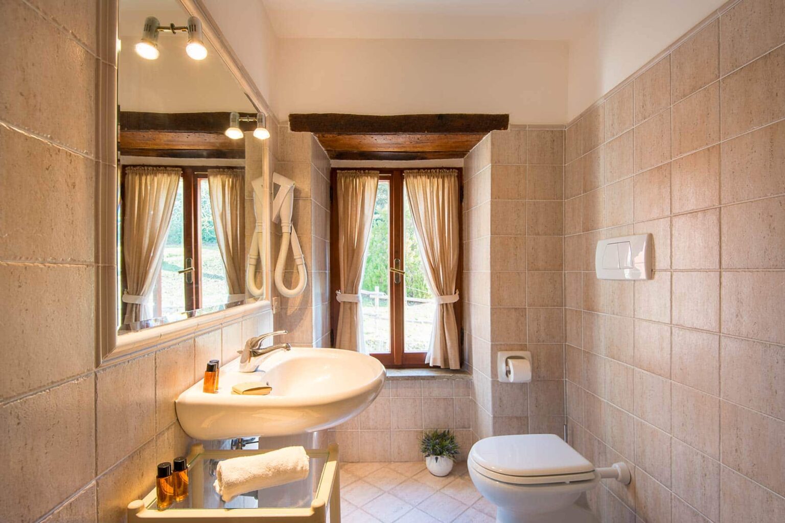 Bright and modern bathroom featuring a sink, mirror, and toilet, with natural light streaming through window curtains, showcasing a clean and inviting design. | Only on SopranoVillas