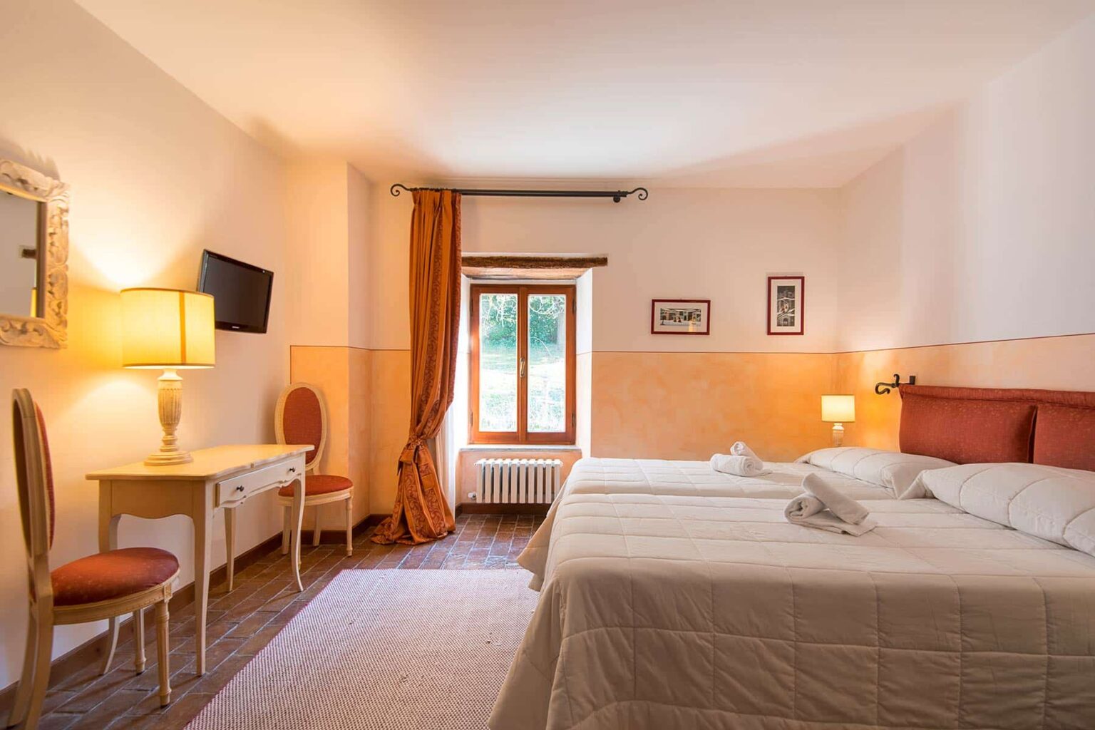 Spacious hotel room featuring two beds, a wooden desk with a chair, a flat-screen TV, and warm lighting. Natural light streams in through a window adorned with curtains, creating a cozy atmosphere. Ideal for travelers seeking comfort and relaxation. | Only on SopranoVillas