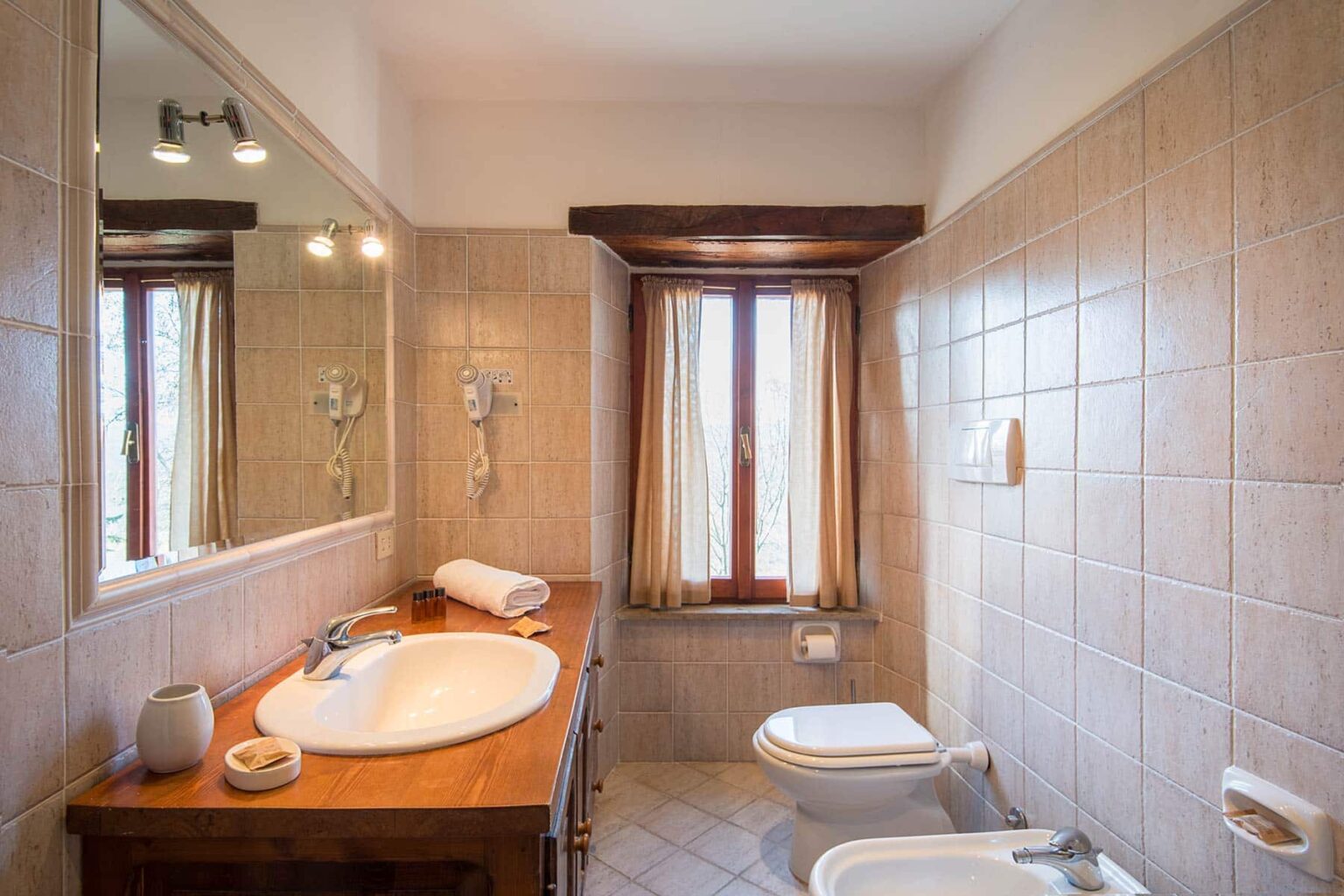 Bright and modern bathroom featuring a large mirror, wooden vanity with sink, and natural light from a window adorned with curtains. The space includes a toilet and decorative towels, showcasing a clean and inviting design. | Only on SopranoVillas