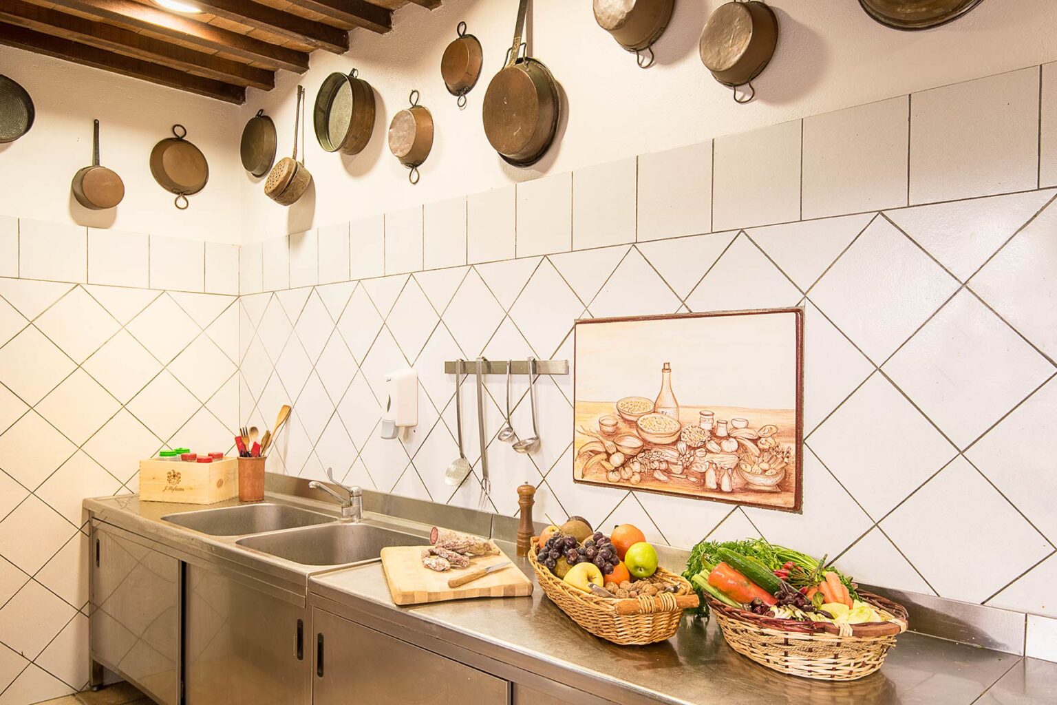Rustic kitchen interior featuring hanging copper pots, a stainless steel sink, and baskets filled with fresh vegetables and fruits on a tiled countertop. A framed artwork depicting a culinary scene adds charm to the space. | Only on SopranoVillas