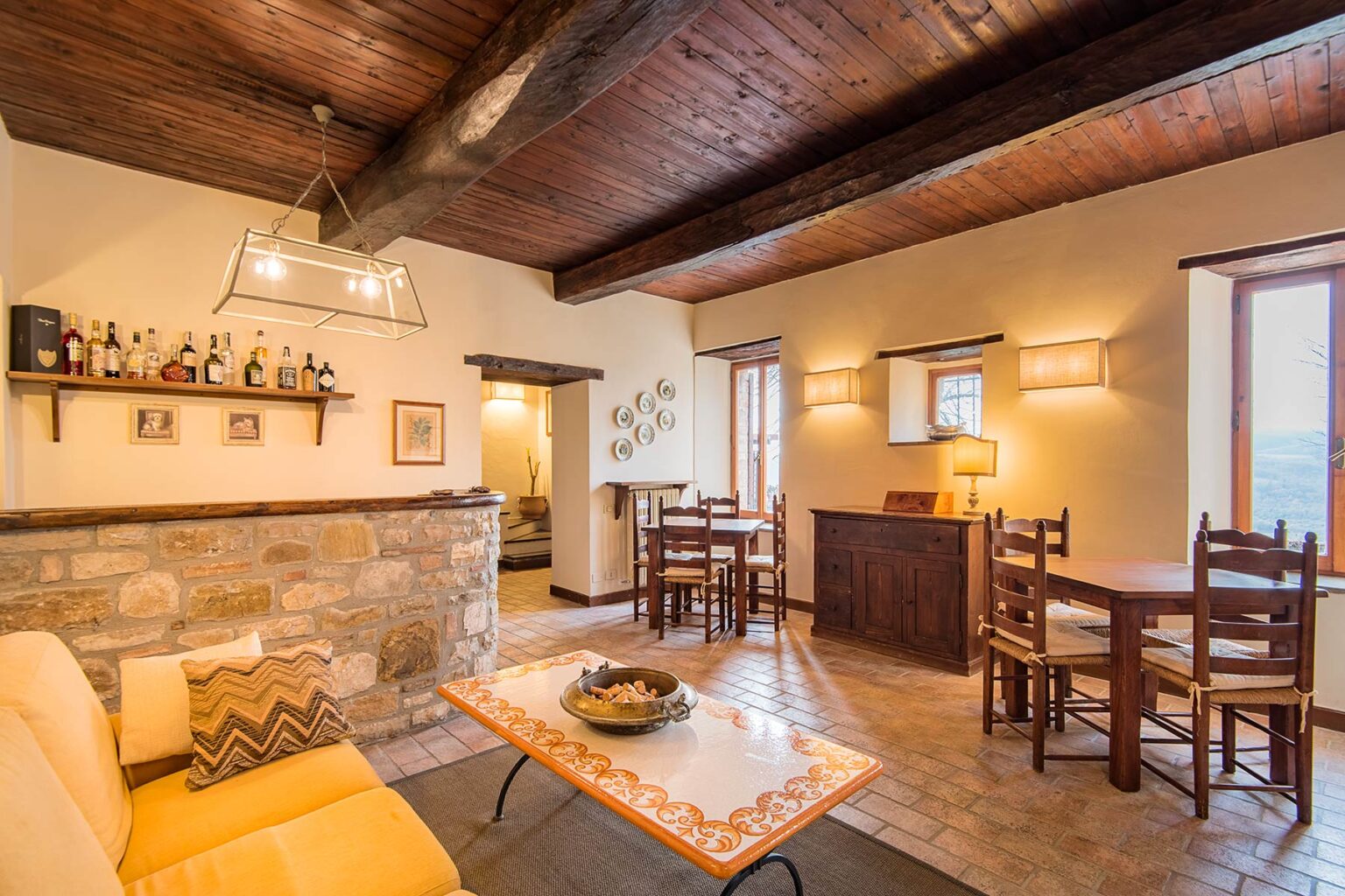 Cozy living room with wooden beams, stone accents, and a warm color palette, featuring a comfortable sofa, a coffee table, and a dining area with wooden chairs. Ideal for relaxation and gatherings. | Only on SopranoVillas