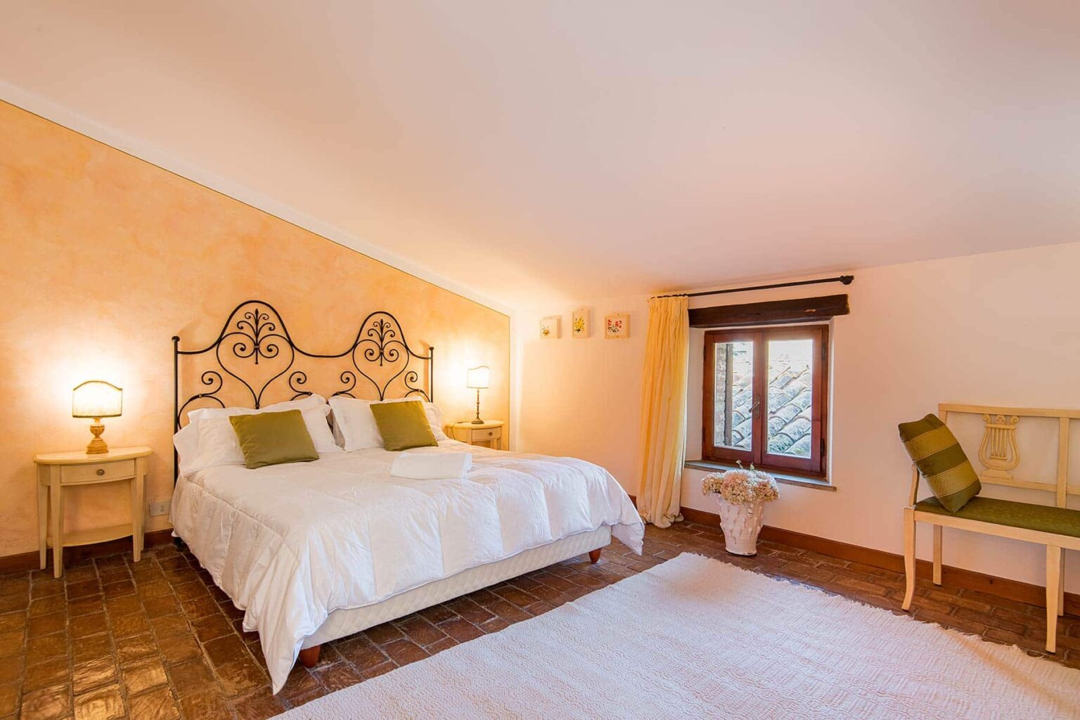 Cozy bedroom featuring a large bed with decorative headboard, soft lighting, and warm peach-colored walls. Includes a window with curtains, a small table, and a chair, creating an inviting atmosphere for relaxation. | Only on SopranoVillas