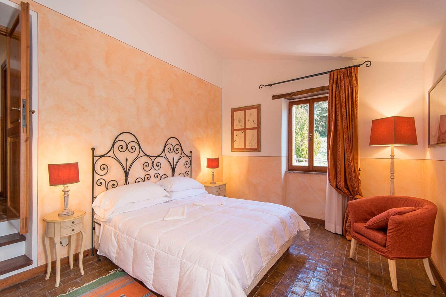 Cozy bedroom featuring a wrought iron bed, warm orange walls, and elegant decor. The room includes bedside lamps, a comfortable armchair, and a window with natural light, creating a welcoming atmosphere for relaxation. | Only on SopranoVillas