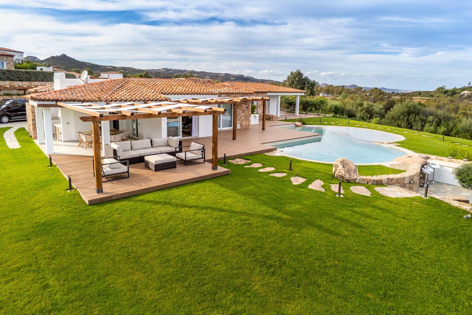 Modern villa with a wooden pergola, outdoor seating area, and a swimming pool surrounded by lush green grass and scenic views. Perfect for luxury living or vacation rentals. | Only on SopranoVillas