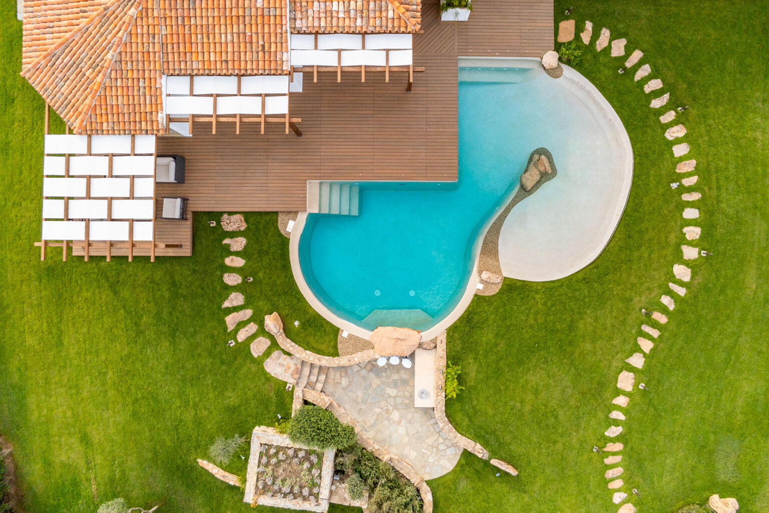 Aerial view of a luxurious villa with a swimming pool, surrounded by landscaped gardens and stone pathways. The property features a wooden deck and a tiled roof, set against a vibrant green lawn. | Only on SopranoVillas