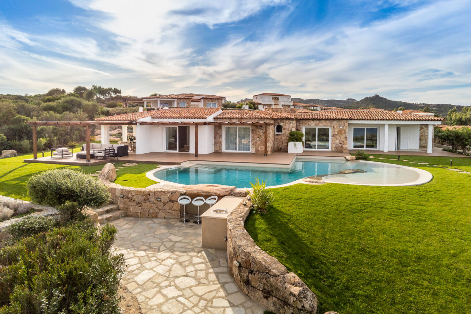 Luxurious villa with a stone exterior, surrounded by lush greenery and a sparkling swimming pool, set against a backdrop of blue skies and rolling hills. Perfect for relaxation and outdoor living. | Only on SopranoVillas