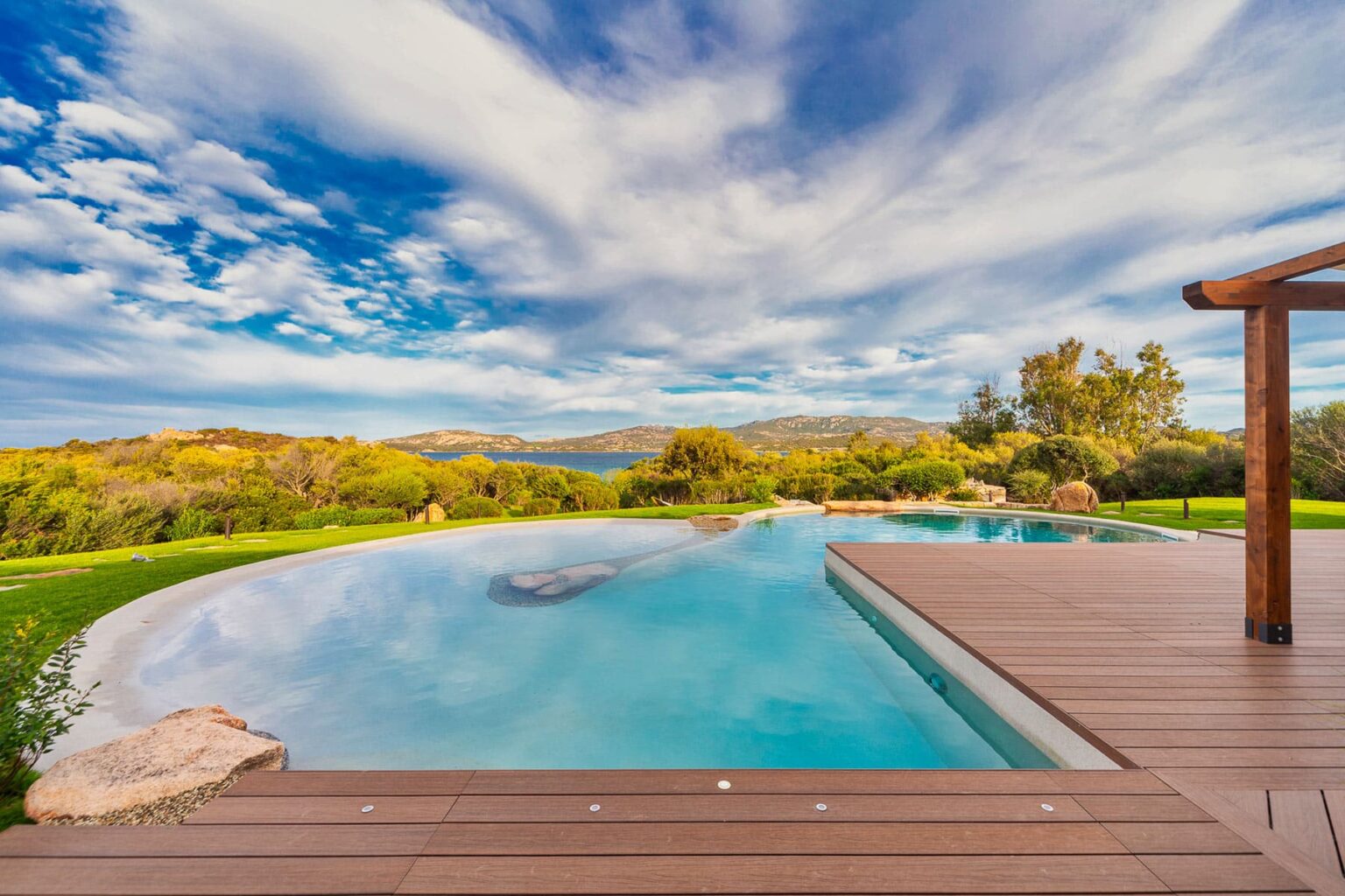 Scenic view of a modern infinity pool surrounded by lush greenery and vibrant landscaping, under a dramatic sky with clouds, showcasing a tranquil outdoor setting. | Only on SopranoVillas