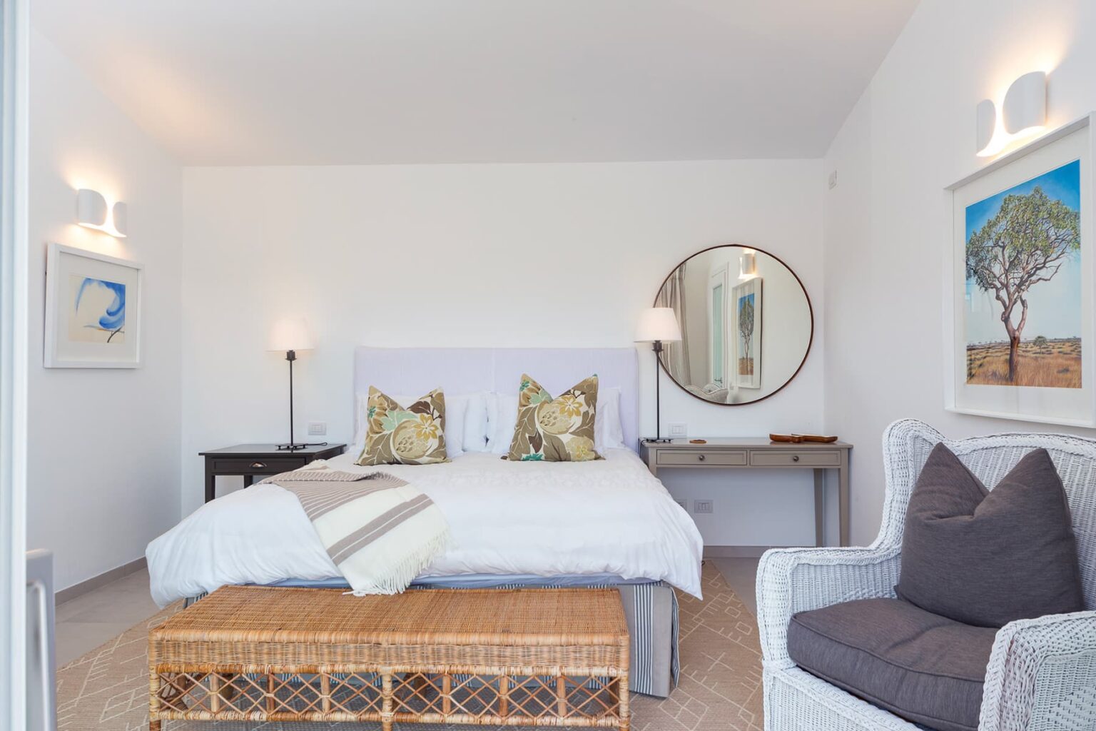 Modern bedroom interior featuring a king-size bed with decorative pillows, a stylish round mirror, and a cozy armchair. Natural light enhances the serene atmosphere, complemented by artwork on the walls and a woven bench at the foot of the bed. Ideal for relaxation and comfort. | Only on SopranoVillas