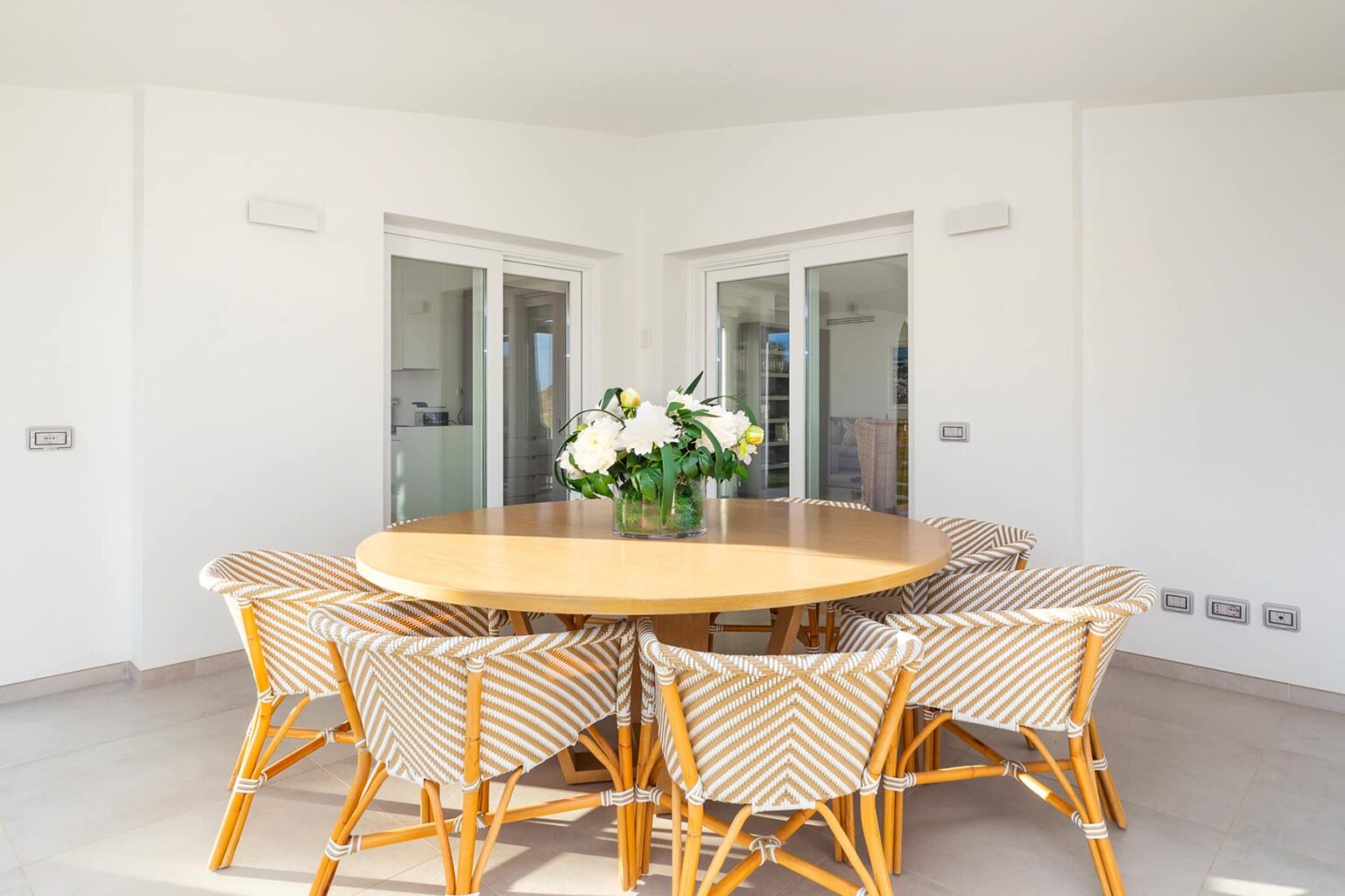 Modern dining area featuring a round wooden table surrounded by stylish woven chairs and a floral centerpiece, with large windows allowing natural light. | Only on SopranoVillas