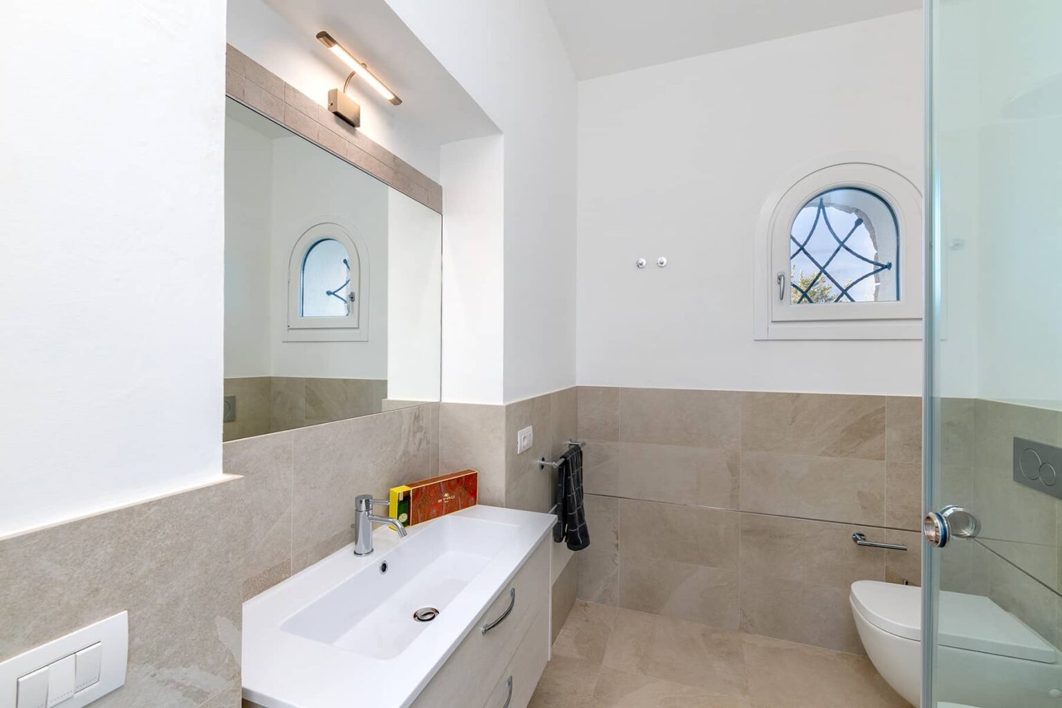 Modern bathroom featuring a sleek vanity with a large mirror, natural stone tiles, and a glass shower enclosure, illuminated by soft lighting and a decorative window. | Only on SopranoVillas