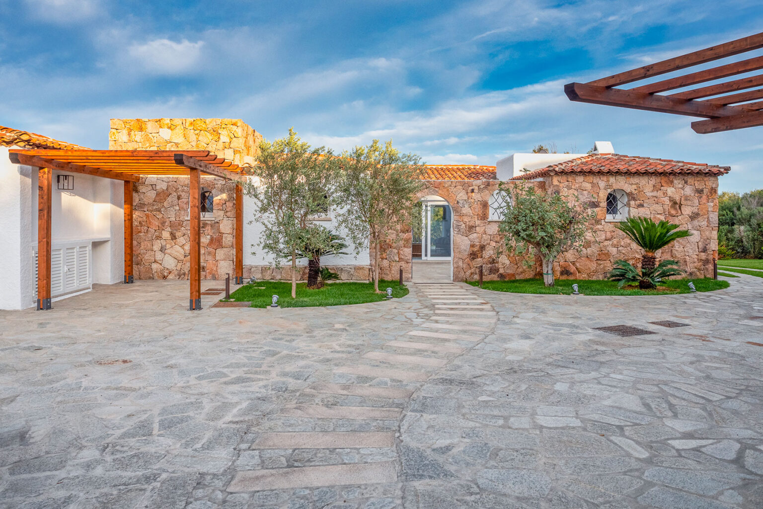 Modern stone villa with a landscaped entrance, featuring olive trees and a stone pathway, set against a blue sky. Ideal for luxury home buyers or vacation rentals. | Only on SopranoVillas
