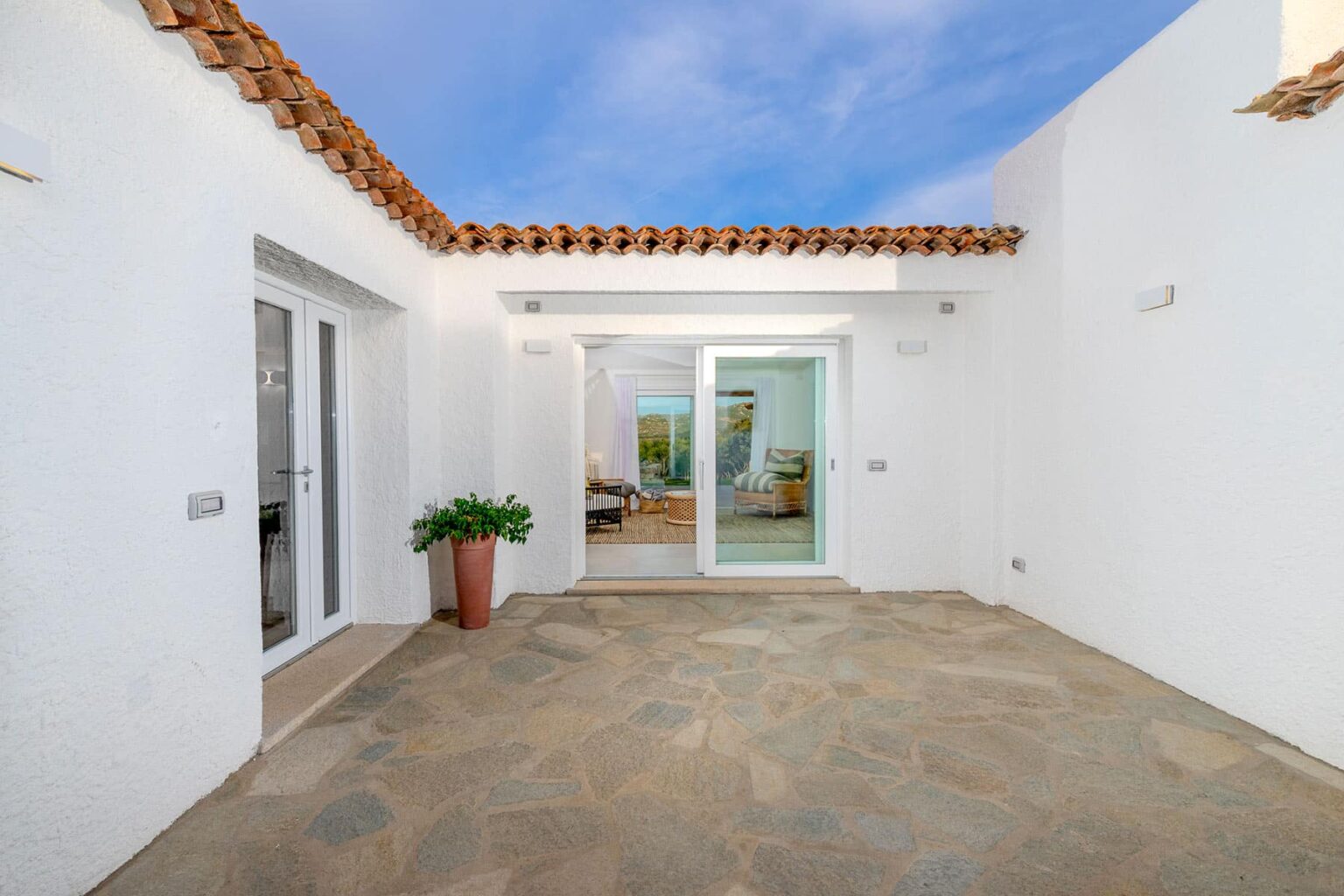 Modern outdoor patio area featuring stone flooring, white walls, and a glass door leading into a bright interior space, with a potted plant enhancing the inviting atmosphere. | Only on SopranoVillas