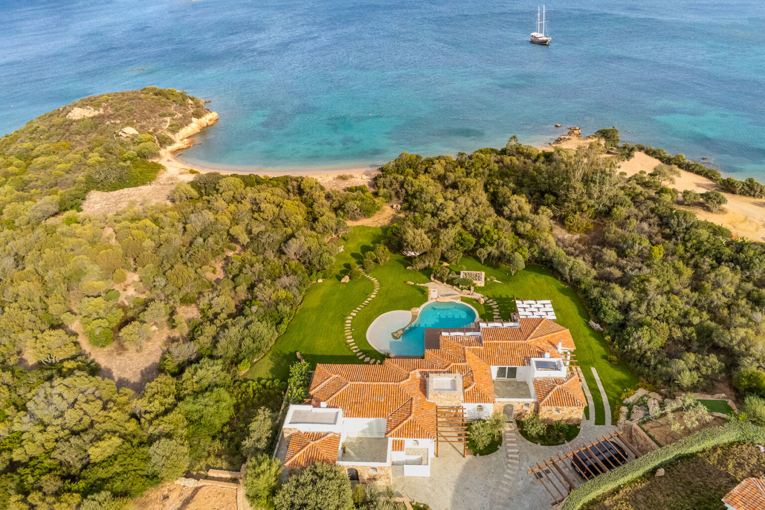 Aerial view of a luxurious coastal villa surrounded by lush greenery, featuring a swimming pool and expansive outdoor space, with a serene beach and ocean in the background. | Only on SopranoVillas