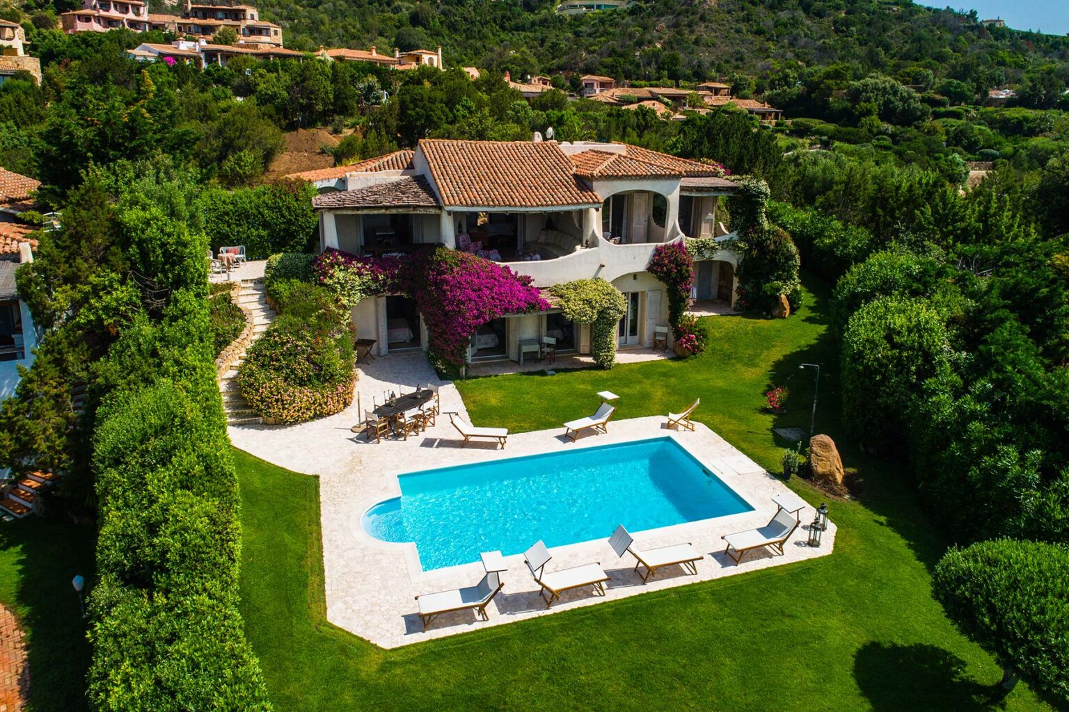 Aerial view of a luxurious villa surrounded by lush greenery, featuring a sparkling blue swimming pool and elegant outdoor seating areas, ideal for relaxation and entertainment. | Only on SopranoVillas