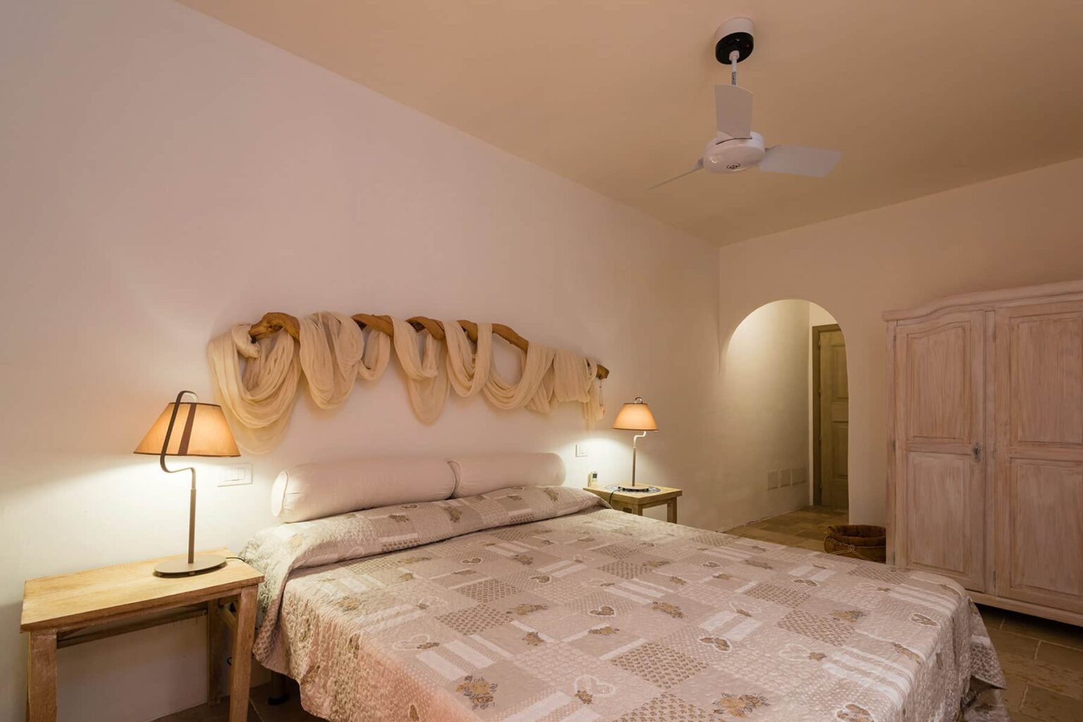 Cozy bedroom featuring a comfortable bed with patterned bedding, soft bedside lamps, and a ceiling fan, complemented by a wooden wardrobe and an arched doorway. Ideal for relaxation and restful nights. | Only on SopranoVillas