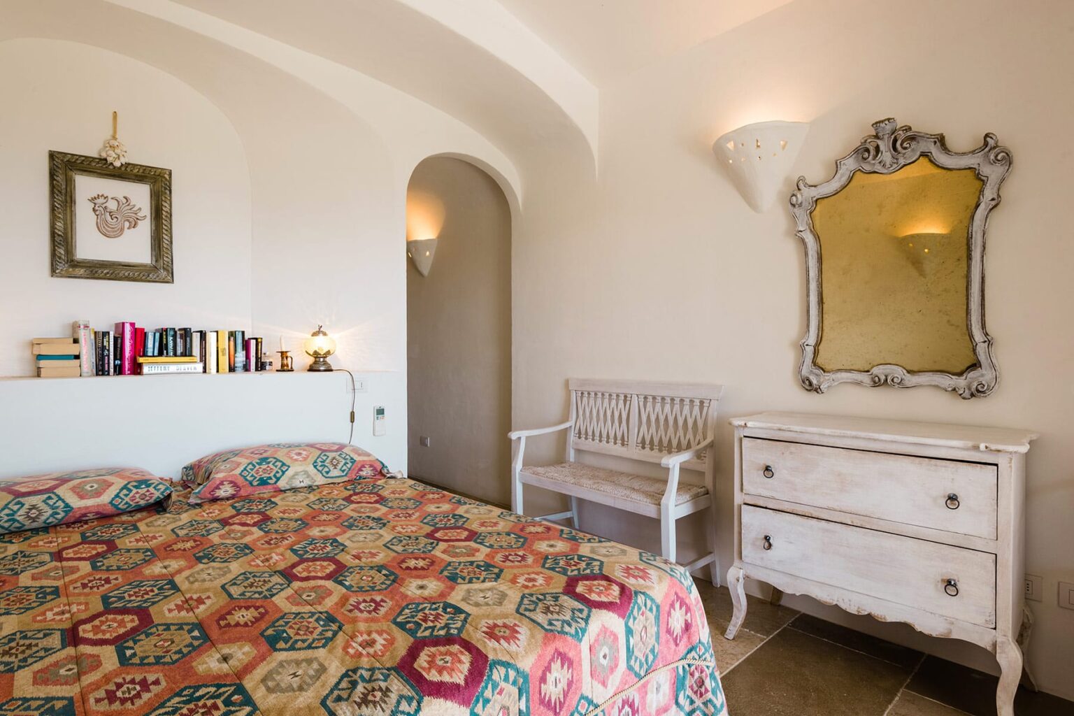 Cozy bedroom featuring a colorful patterned bedspread, vintage-style furniture, an elegant mirror, and a bookshelf, set in a softly lit, inviting atmosphere. | Only on SopranoVillas