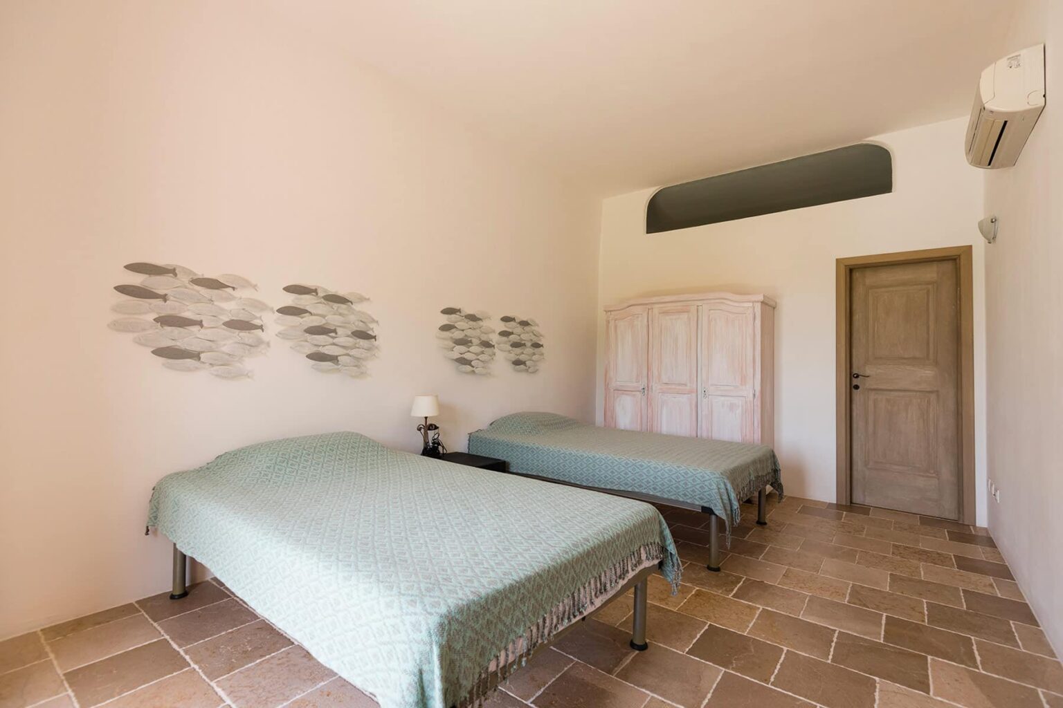 Cozy bedroom featuring two twin beds with light blue bedspreads, decorative fish wall art, and a wooden door. The room has natural stone flooring and an air conditioning unit, creating a comfortable atmosphere. | Only on SopranoVillas