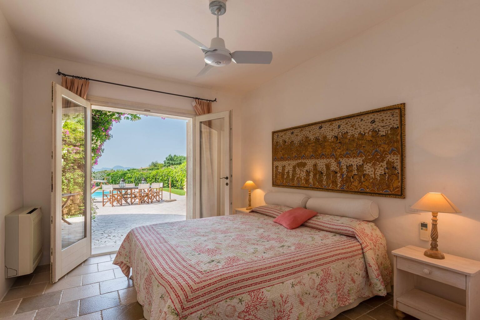 Cozy bedroom with a floral-patterned bedspread, featuring a ceiling fan and bedside lamps. The room opens to a patio with views of a pool and garden, creating a serene indoor-outdoor living space. | Only on SopranoVillas