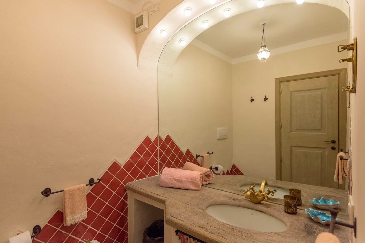 Modern bathroom interior featuring a marble countertop with two sinks, decorative red tile accents, a large mirror with soft lighting, and an elegant wooden door. | Only on SopranoVillas