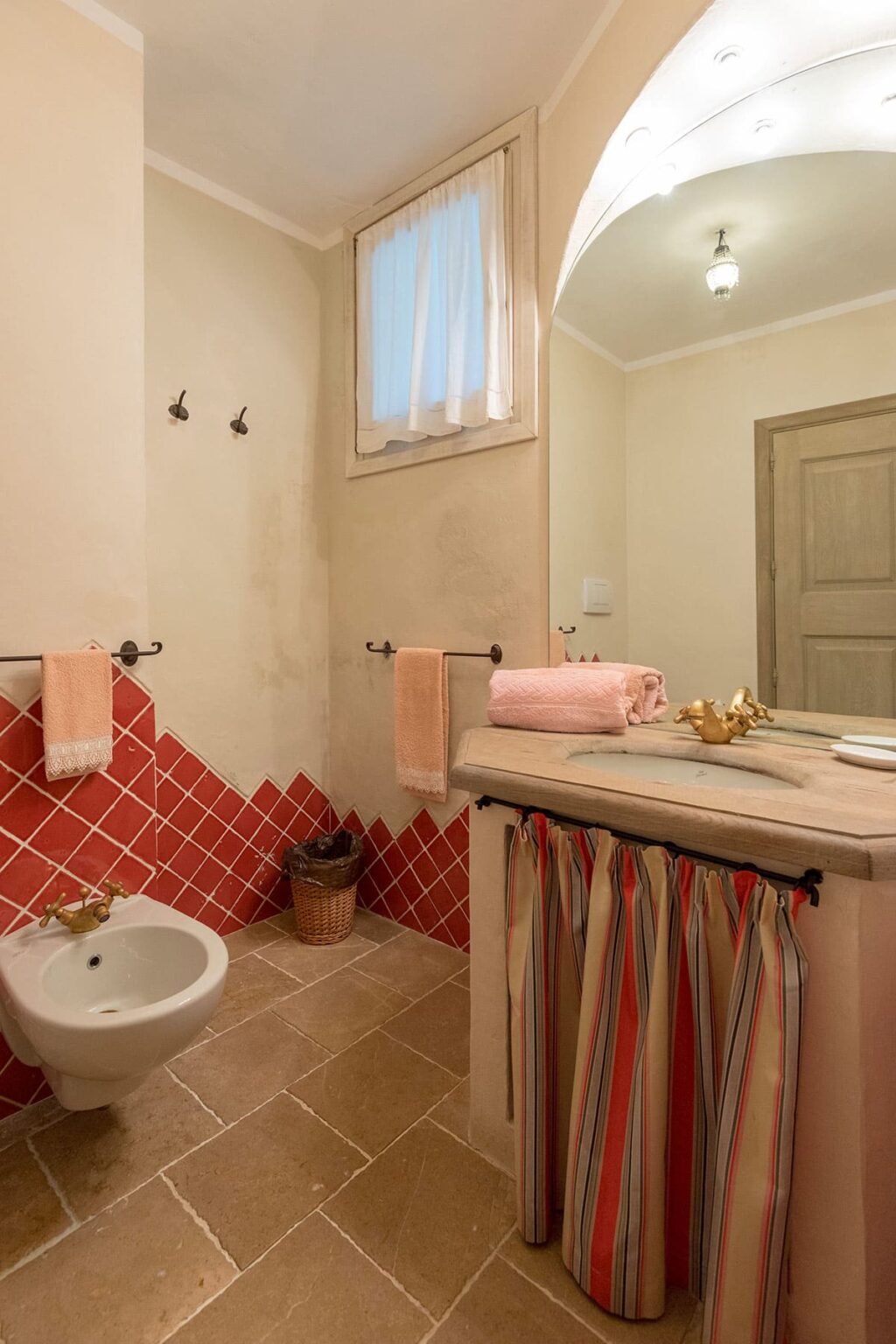 Modern bathroom featuring a decorative red tile wall, a stylish sink with a neutral countertop, and neatly arranged pink towels. The space is well-lit with a window, showcasing a blend of contemporary and rustic design elements. | Only on SopranoVillas