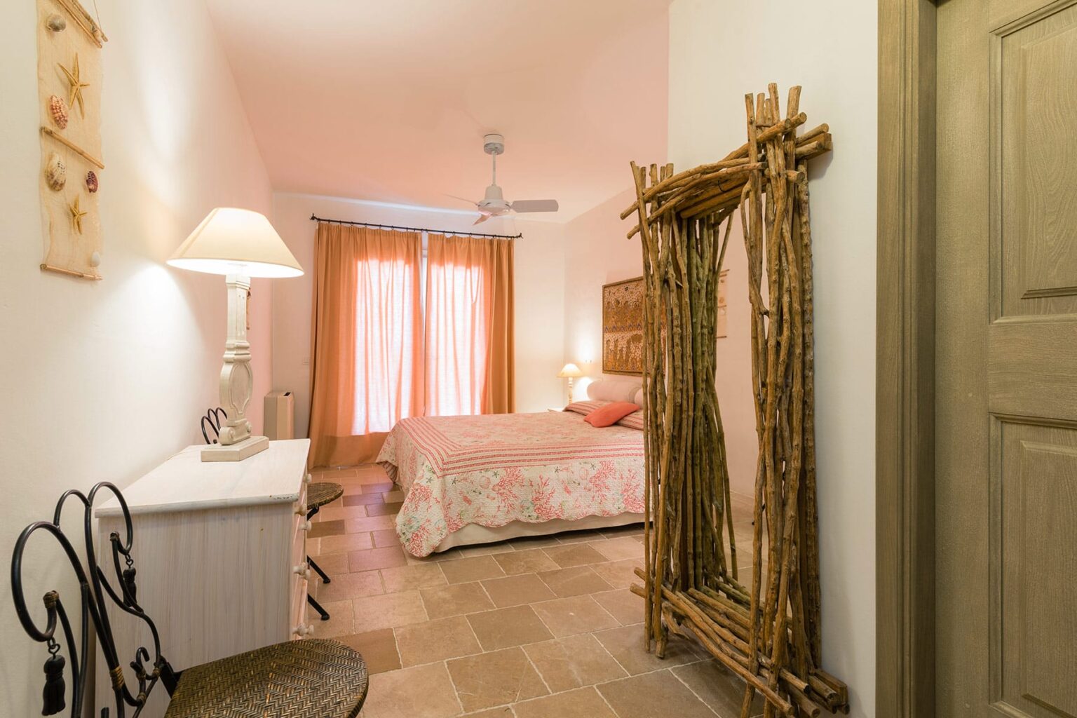 Cozy bedroom featuring a king-sized bed with floral bedding, warm lighting from a bedside lamp, and large windows with sheer curtains. The room includes a rustic wooden mirror and a stylish dresser, creating a welcoming atmosphere. | Only on SopranoVillas