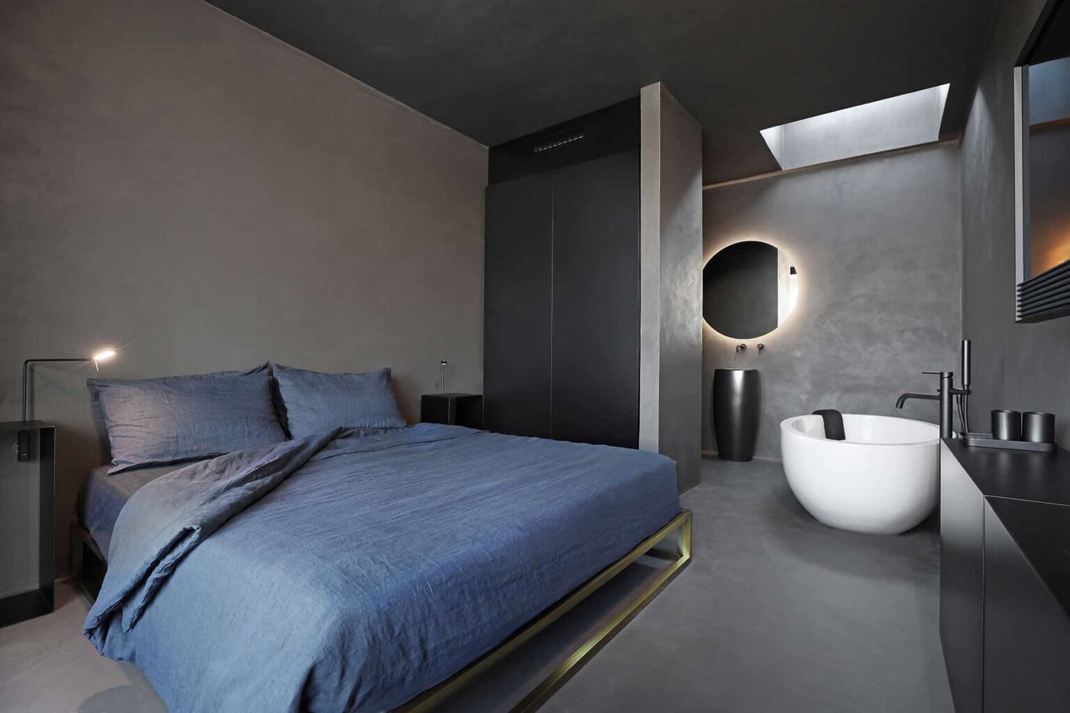 Modern minimalist bedroom featuring a cozy bed with blue bedding, a circular mirror, and a freestanding bathtub. The room has a sleek design with gray walls and natural light from a skylight. Ideal for contemporary interior design inspiration. | Only on SopranoVillas