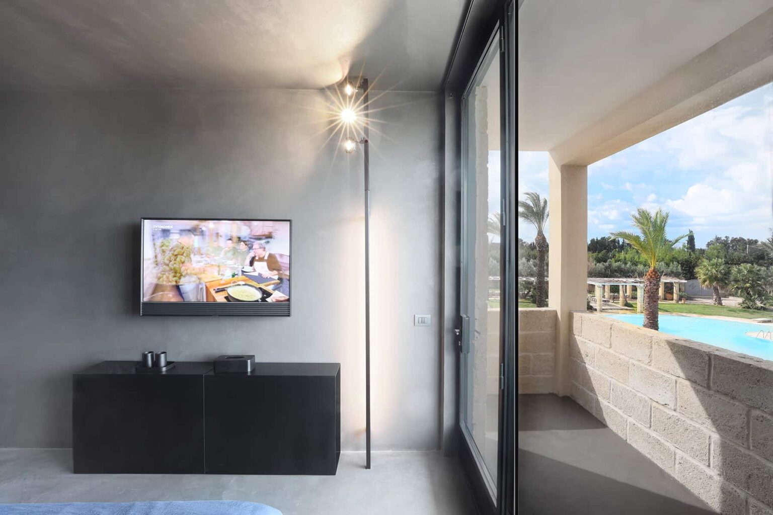 Modern living room with a flat-screen TV and stylish black furniture, featuring large windows that open to a balcony with a view of a pool and palm trees. | Only on SopranoVillas