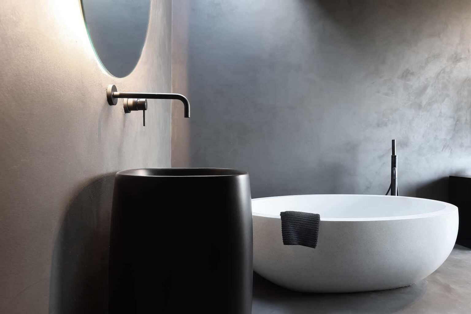 Modern bathroom interior featuring a sleek black faucet, a minimalist round mirror, a stylish black basin, and a contemporary white freestanding bathtub against a textured gray wall. | Only on SopranoVillas
