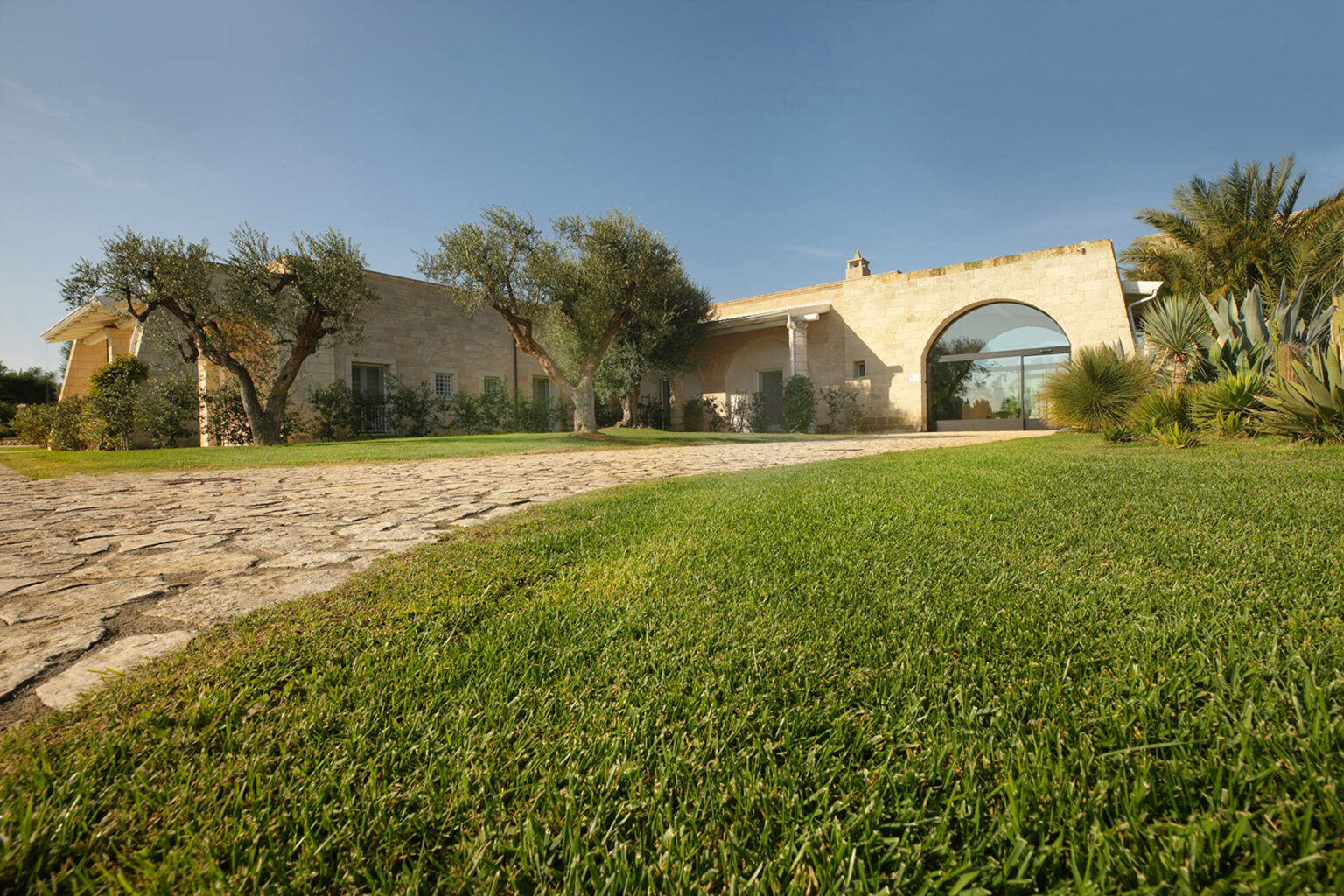 Modern stone villa with an arched entrance, surrounded by lush greenery and palm trees, set against a clear blue sky. The image showcases a well-maintained lawn and a winding stone pathway leading to the entrance. | Only on SopranoVillas