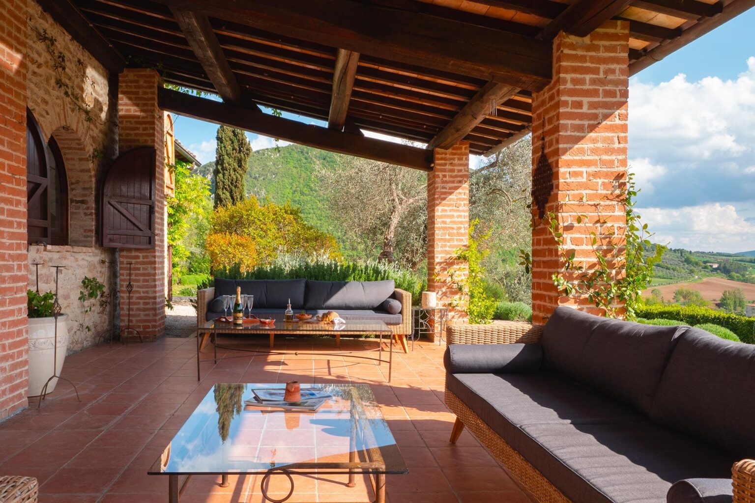 Spacious outdoor patio with brick columns, comfortable seating, and a glass coffee table, surrounded by lush greenery and scenic views of the countryside. | Only on SopranoVillas