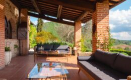 Spacious outdoor patio with brick columns, comfortable seating, and a glass coffee table, surrounded by lush greenery and scenic views of the countryside.