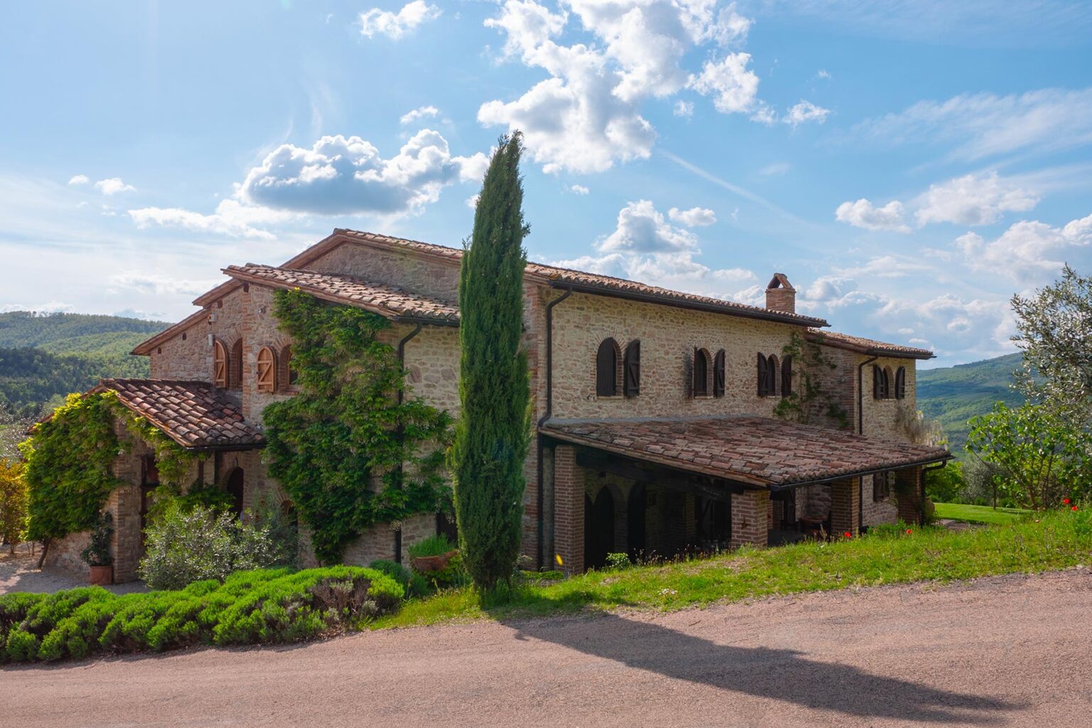 Historic stone villa surrounded by lush greenery and cypress trees, set against a clear blue sky in a picturesque landscape. Ideal for travelers seeking rustic charm in Tuscany. | Only on SopranoVillas