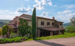 Historic stone villa surrounded by lush greenery and cypress trees, set against a clear blue sky in a picturesque landscape. Ideal for travelers seeking rustic charm in Tuscany.