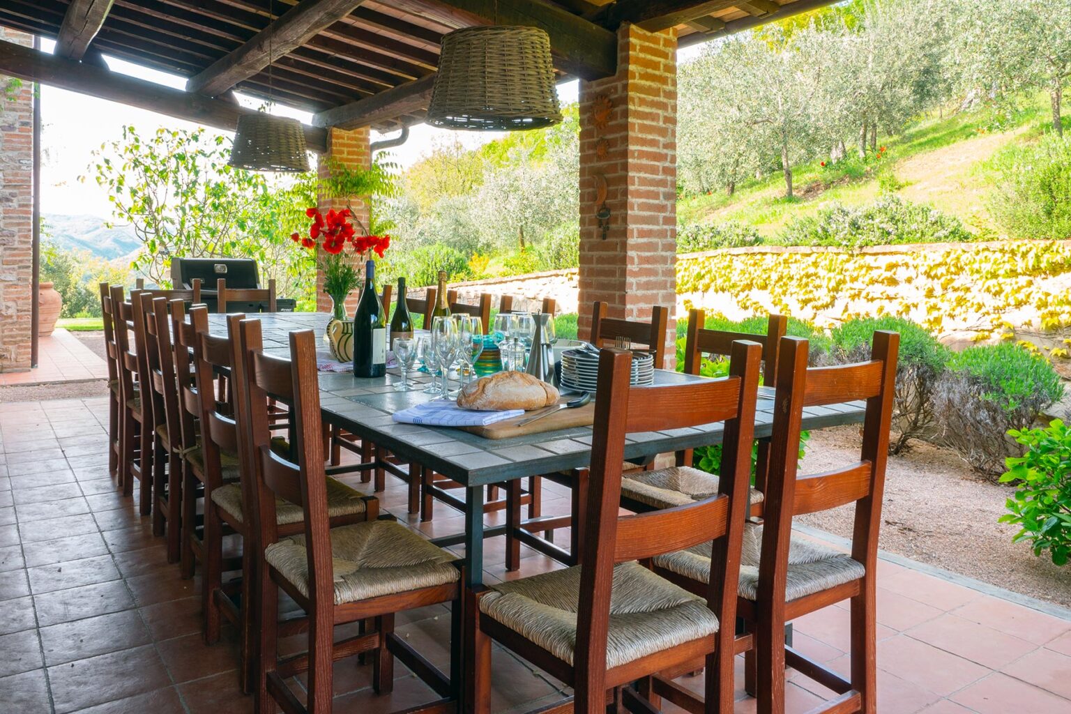 Outdoor dining area with a long wooden table set for a meal, surrounded by chairs, featuring glasses, wine bottles, and a loaf of bread, with a scenic view of green hills and olive trees in the background. Perfect for gatherings and al fresco dining experiences. | Only on SopranoVillas