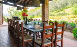 Outdoor dining area with a long wooden table set for a meal, surrounded by chairs, featuring glasses, wine bottles, and a loaf of bread, with a scenic view of green hills and olive trees in the background. Perfect for gatherings and al fresco dining experiences.