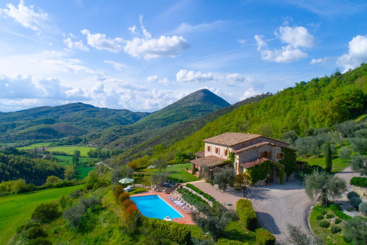 Aerial view of a picturesque villa with a swimming pool surrounded by lush green hills and mountains under a blue sky with fluffy clouds. Ideal for luxury vacation rentals in a serene countryside setting. | Only on SopranoVillas