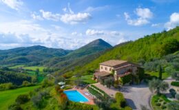 Aerial view of a picturesque villa with a swimming pool surrounded by lush green hills and mountains under a blue sky with fluffy clouds. Ideal for luxury vacation rentals in a serene countryside setting.