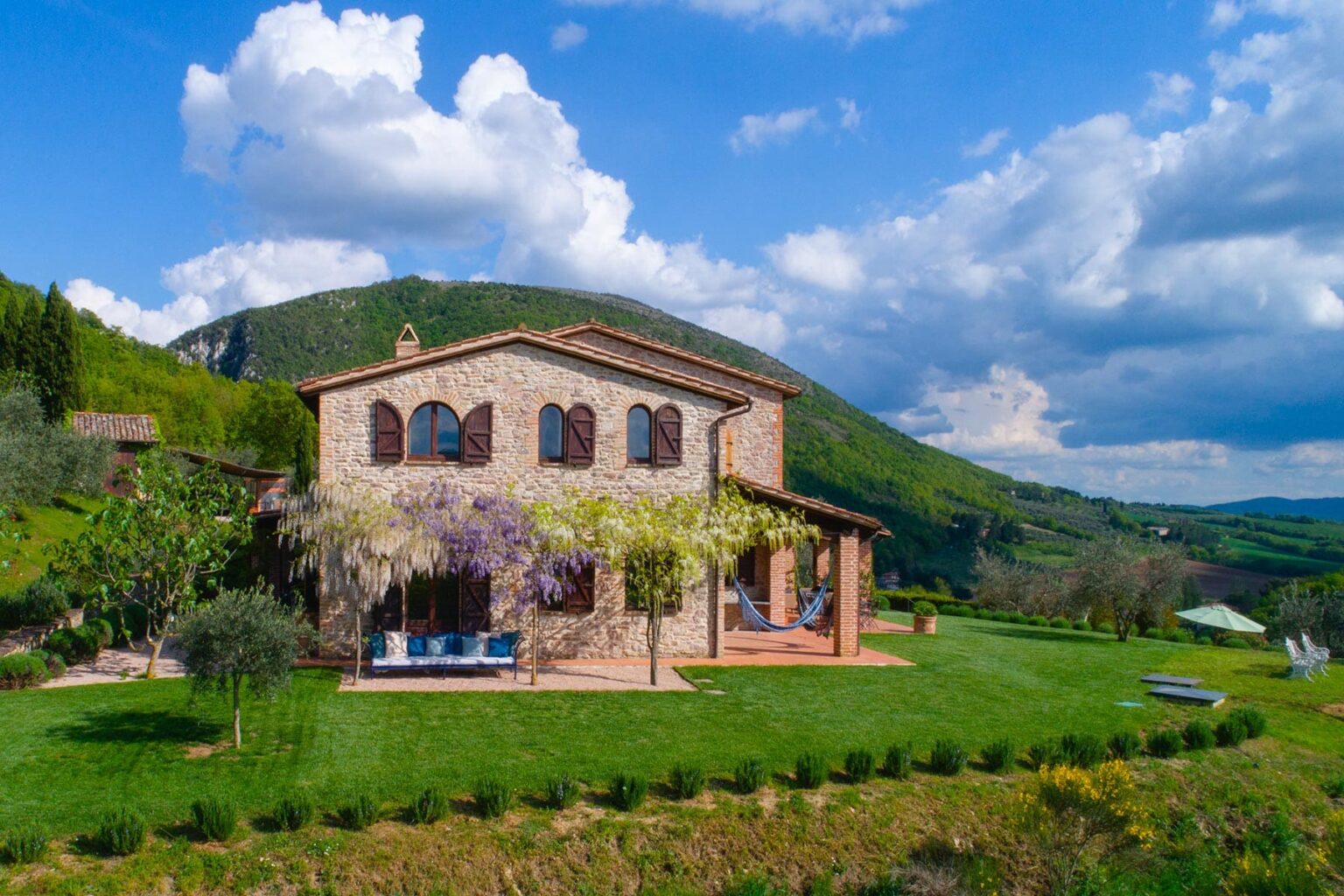 Scenic view of a rustic stone villa surrounded by lush greenery and rolling hills under a blue sky with fluffy clouds, ideal for vacation rentals or countryside retreats. | Only on SopranoVillas