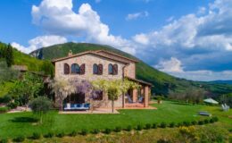 Scenic view of a rustic stone villa surrounded by lush greenery and rolling hills under a blue sky with fluffy clouds, ideal for vacation rentals or countryside retreats.