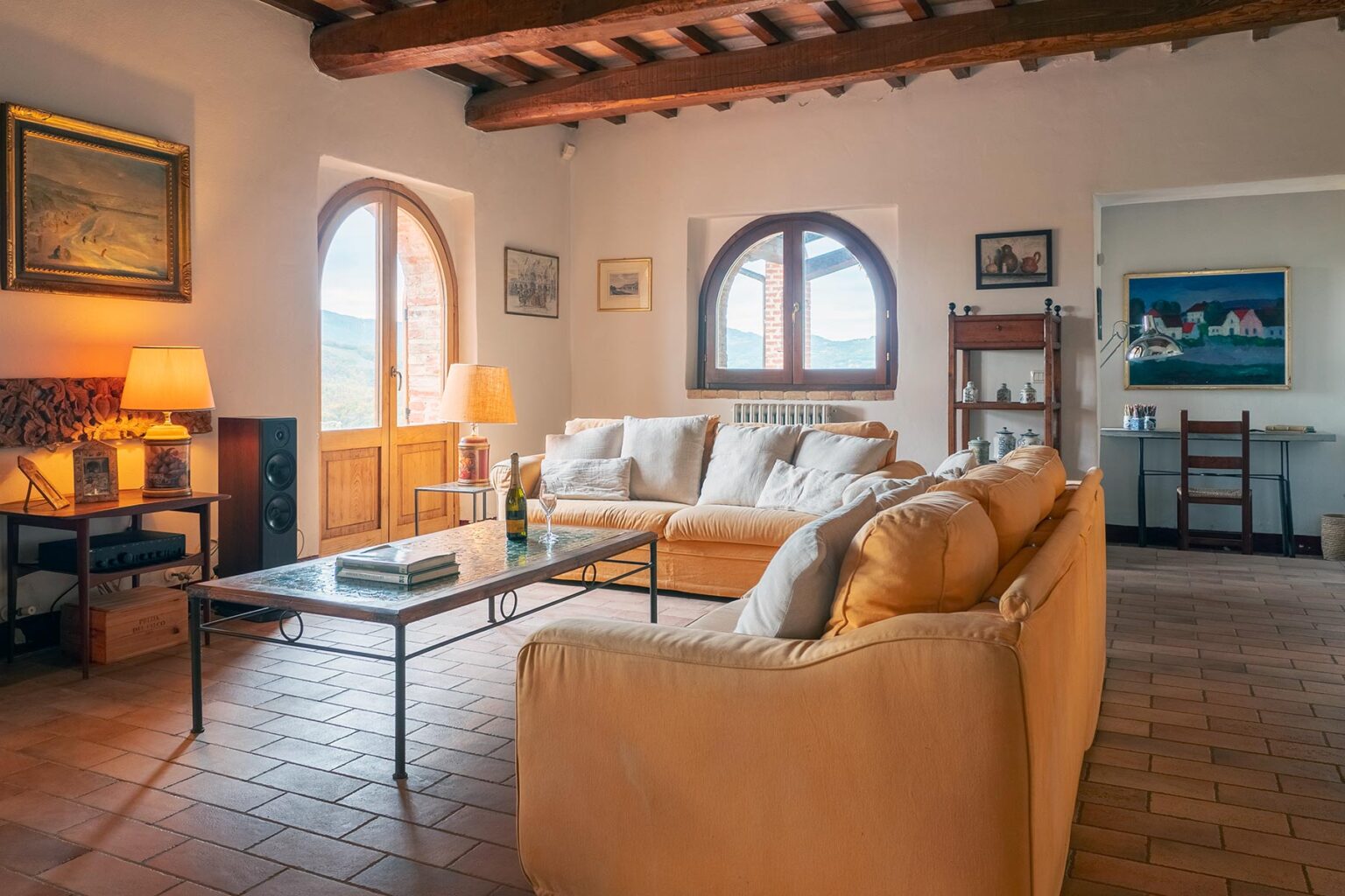 Spacious living room with rustic decor, featuring a comfortable beige sofa, a coffee table, and large windows that allow natural light. The room has wooden beams and artistic wall decor, creating a cozy atmosphere. | Only on SopranoVillas
