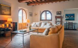 Spacious living room with rustic decor, featuring a comfortable beige sofa, a coffee table, and large windows that allow natural light. The room has wooden beams and artistic wall decor, creating a cozy atmosphere.