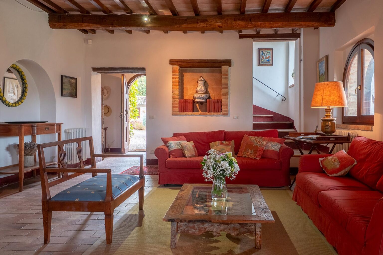 Cozy living room featuring red sofas, wooden furniture, and decorative artwork. A glass coffee table sits in the center, complemented by a vase of flowers. Natural light streams through arched windows, enhancing the warm ambiance. | Only on SopranoVillas