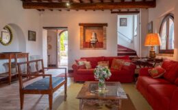 Cozy living room featuring red sofas, wooden furniture, and decorative artwork. A glass coffee table sits in the center, complemented by a vase of flowers. Natural light streams through arched windows, enhancing the warm ambiance.