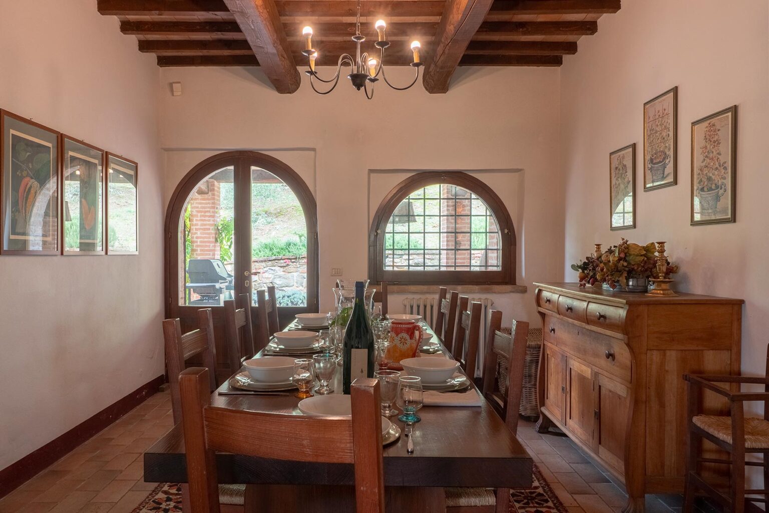 Cozy dining room featuring a long wooden table set for a meal, with rustic chairs, decorative wall art, and large arched windows that provide natural light. A wooden sideboard displays elegant tableware, enhancing the inviting atmosphere. | Only on SopranoVillas