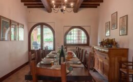 Cozy dining room featuring a long wooden table set for a meal, with rustic chairs, decorative wall art, and large arched windows that provide natural light. A wooden sideboard displays elegant tableware, enhancing the inviting atmosphere.