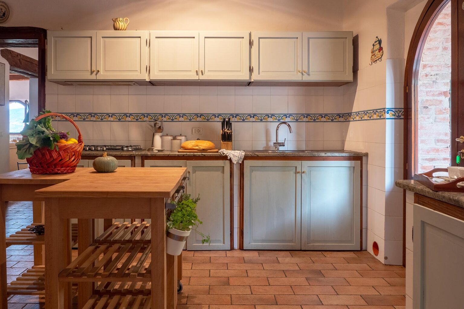 Cozy kitchen interior featuring light green cabinets, a wooden island, and tiled backsplash, with fresh produce in a basket and a bright, inviting atmosphere. | Only on SopranoVillas